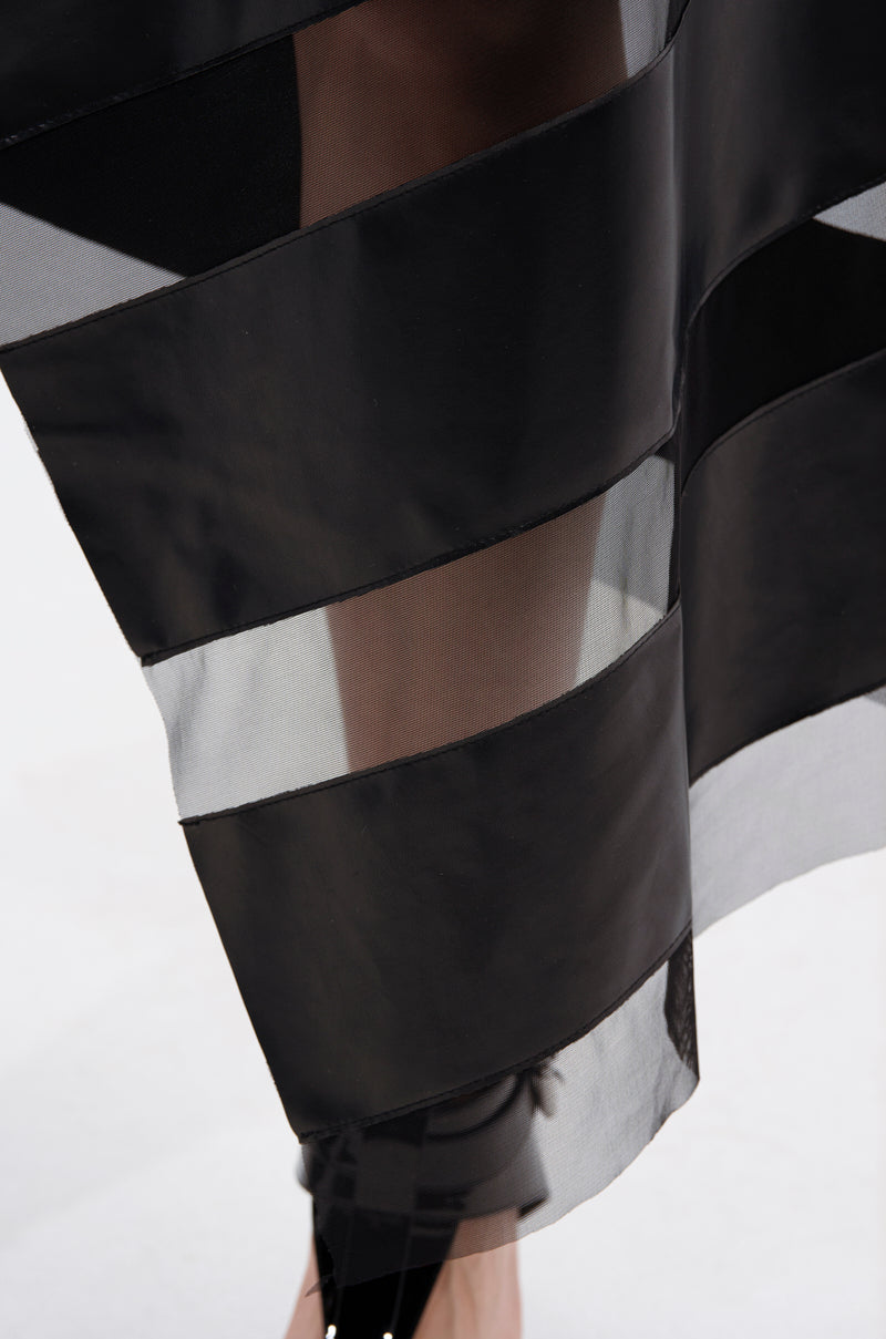 Close-up of the SOMETHING DIFFERENT MESH MIDI SKIRT WITH FAUX LEATHER, featuring alternating sheer and opaque black stripes that reveal part of a leg and black shoe against a plain light background.