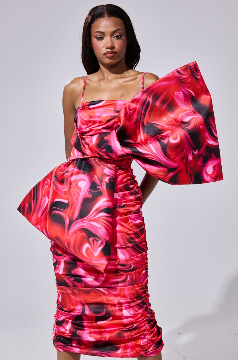 A woman wears the ONE OF ONE BOW MIDI DRESS, a vibrant satin piece with an asymmetrical silhouette, swirling red, pink, and black patterns, and dramatic sculptural ruffles on one shoulder and hip. She stands against a plain light gray background.