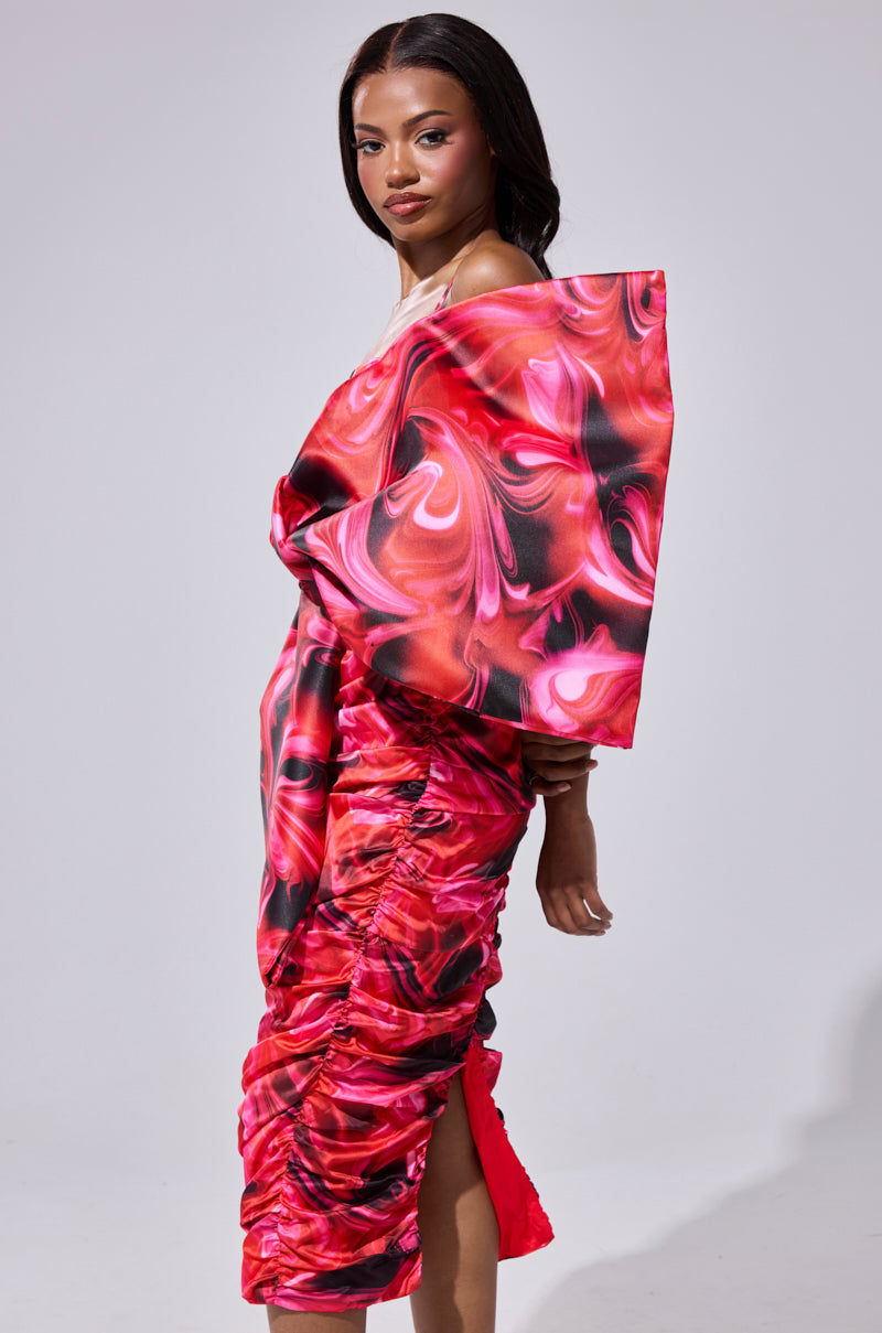 A woman poses confidently in the ONE OF ONE BOW MIDI DRESS, a bold pink and red piece with swirling patterns, sculptural shoulders, and ruched details, standing against a plain gray background.