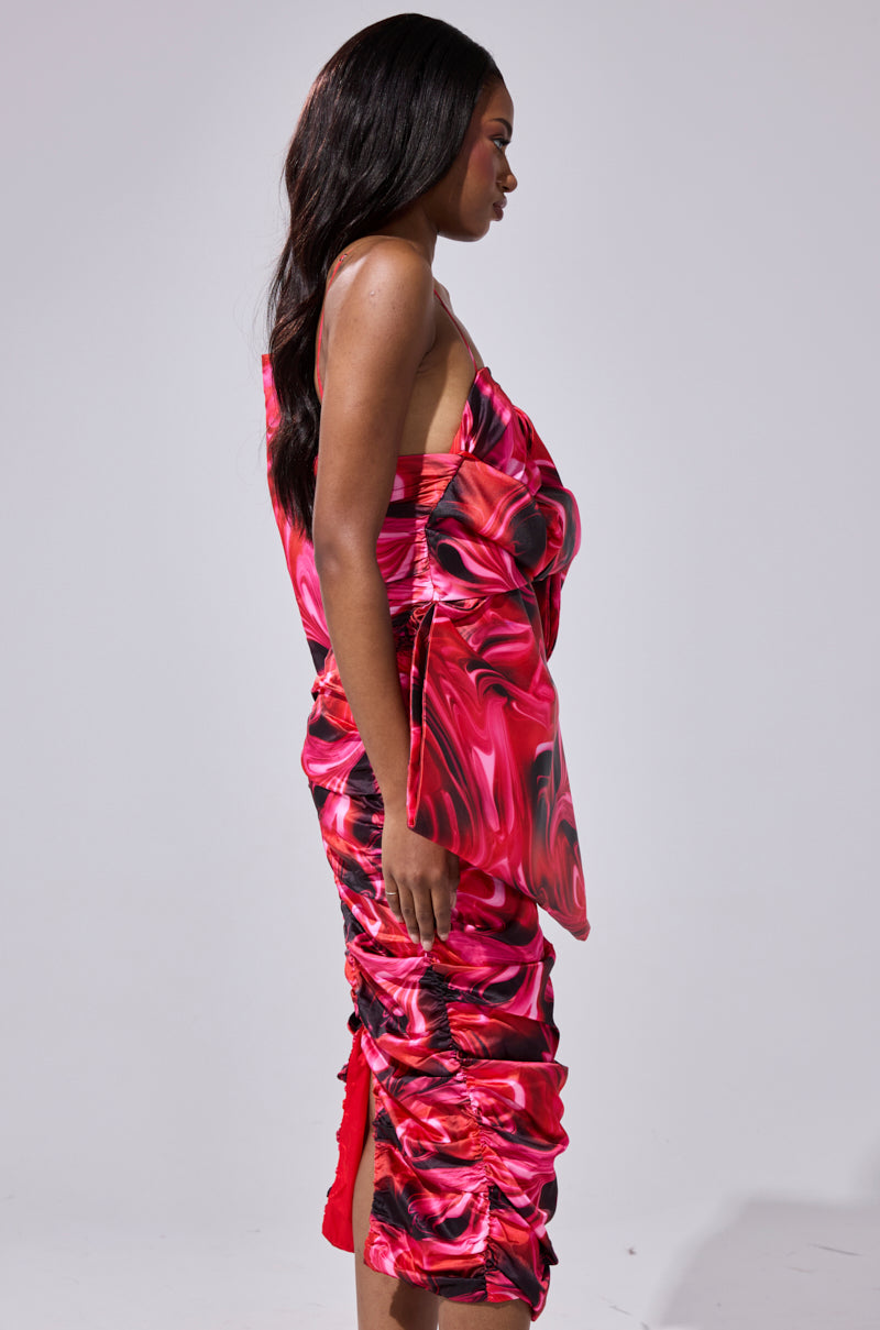 A woman in profile wears the ONE OF ONE BOW MIDI DRESS, featuring vibrant red and pink swirls, ruched bodycon fit, draped asymmetrical design. She has long dark hair and a neutral expression against a plain light background.