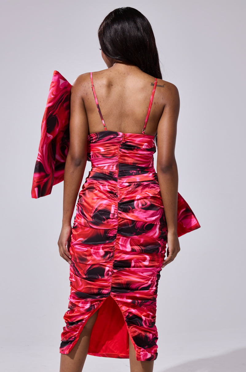 A woman faces away, wearing the ONE OF ONE BOW MIDI DRESS—a ruched bodycon with bold red and black abstract print, spaghetti straps, and a dramatic asymmetrical bow. Her long dark hair cascades down her back, radiating high-fashion drama.