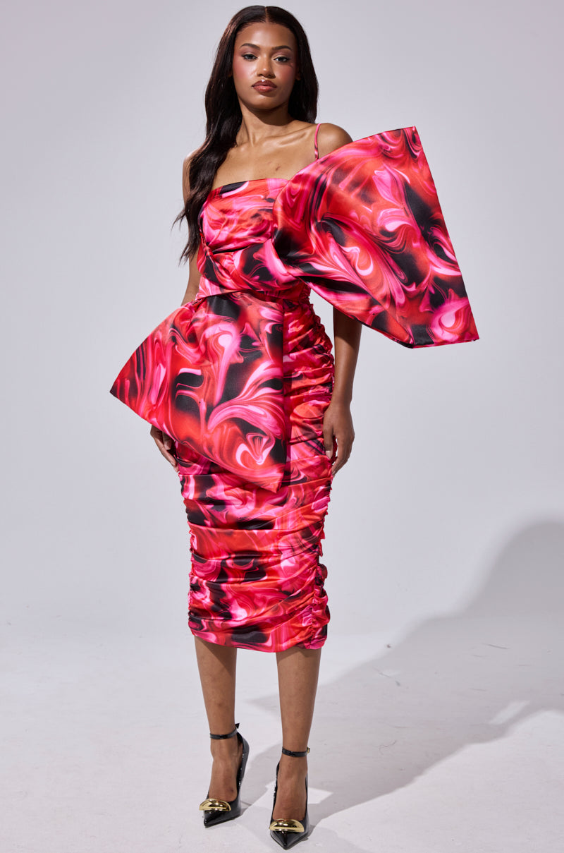 A woman models the ONE OF ONE BOW MIDI DRESS, a bold satin bodycon with a vibrant red and pink marble pattern, an exaggerated single sleeve, and sculptural side detail. She pairs it with metallic pointed heels against a plain light background.