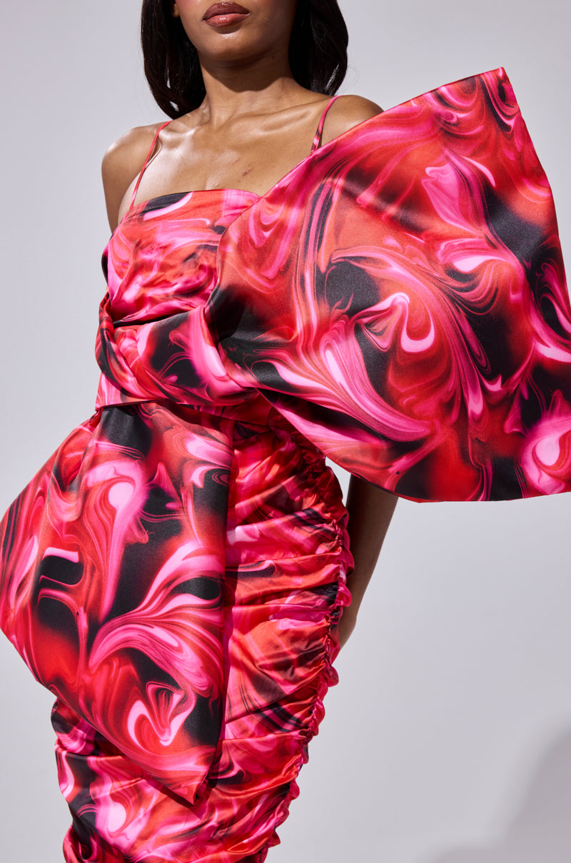 A woman wears the ONE OF ONE BOW MIDI DRESS featuring a vibrant abstract pattern in pink, red, and black, accented with oversized sculpted details at the shoulder and hip. She stands against a plain, light background.