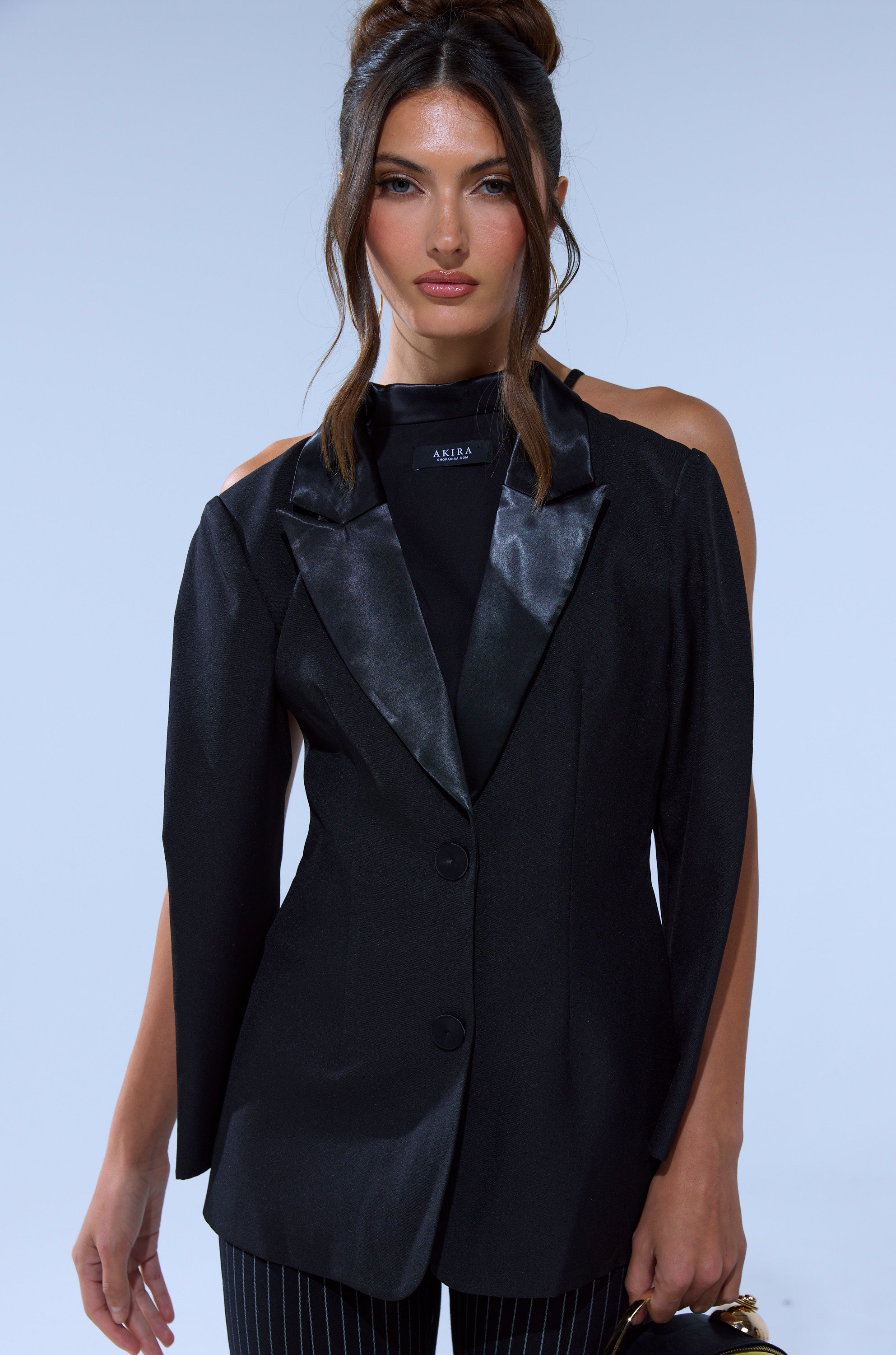 A woman with long brown hair in a bun wears the TELL ME ABOUT IT BUTTON DOWN BLAZER BLOUSE over a black top, paired with pinstriped pants, standing against a light blue background.