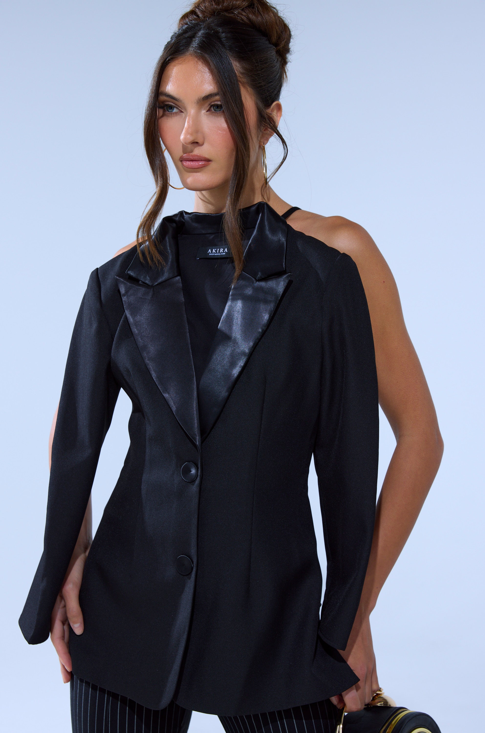 A woman wears the TELL ME ABOUT IT BUTTON DOWN BLAZER BLOUSE—black with one satin lapel, one full sleeve, and one sleeveless side—paired with pinstripe pants. Her long brown hair is in an updo as she carries a small bag.