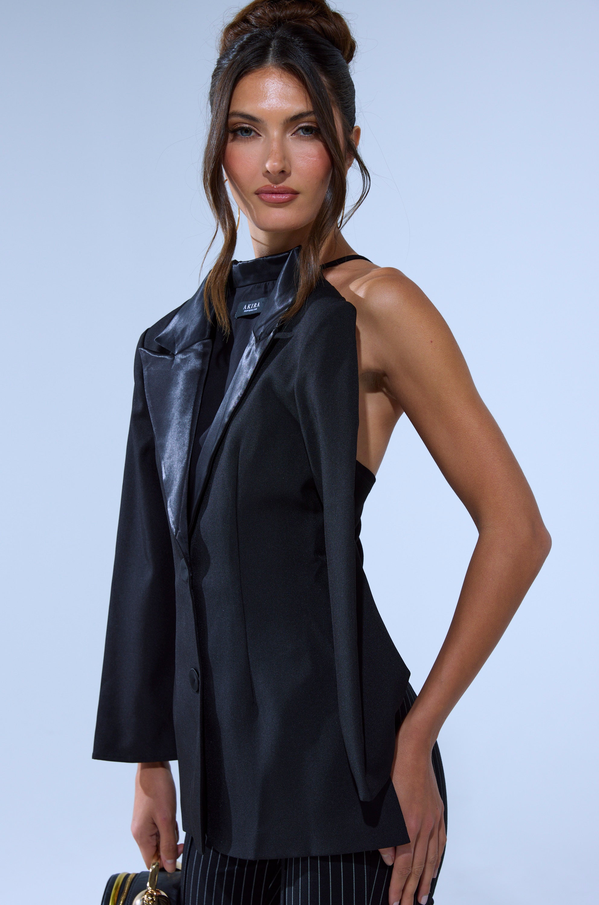 A woman poses confidently in the TELL ME ABOUT IT BUTTON DOWN BLAZER BLOUSE, a unique black one-sleeve design that bares her shoulder and arm. She wears pinstripe pants, with her long brown hair styled up against a light blue background.