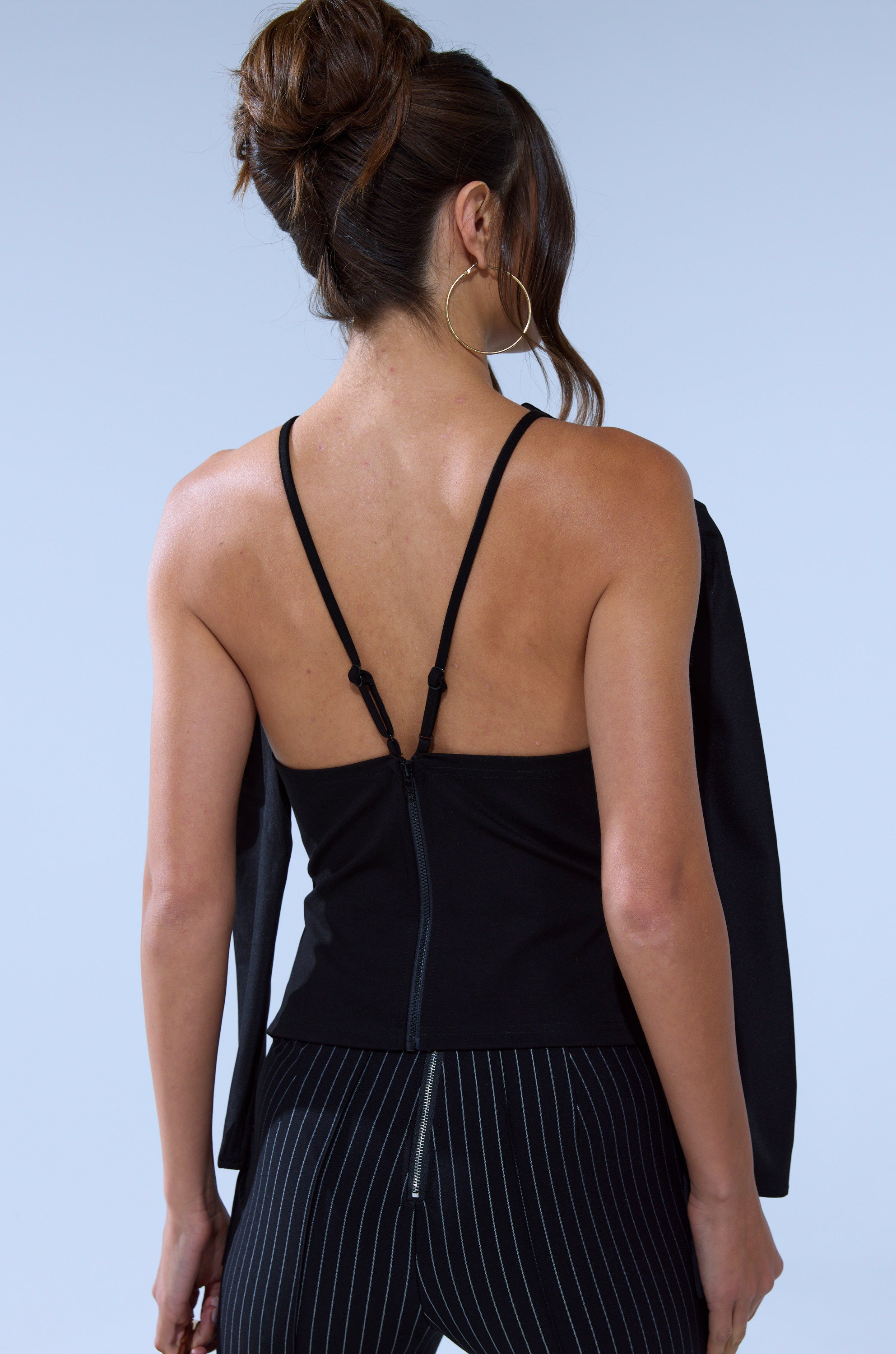 A woman with her hair in a bun, wearing the TELL ME ABOUT IT BUTTON DOWN BLAZER BLOUSE and black pinstriped pants, is facing away from the camera against a light background.