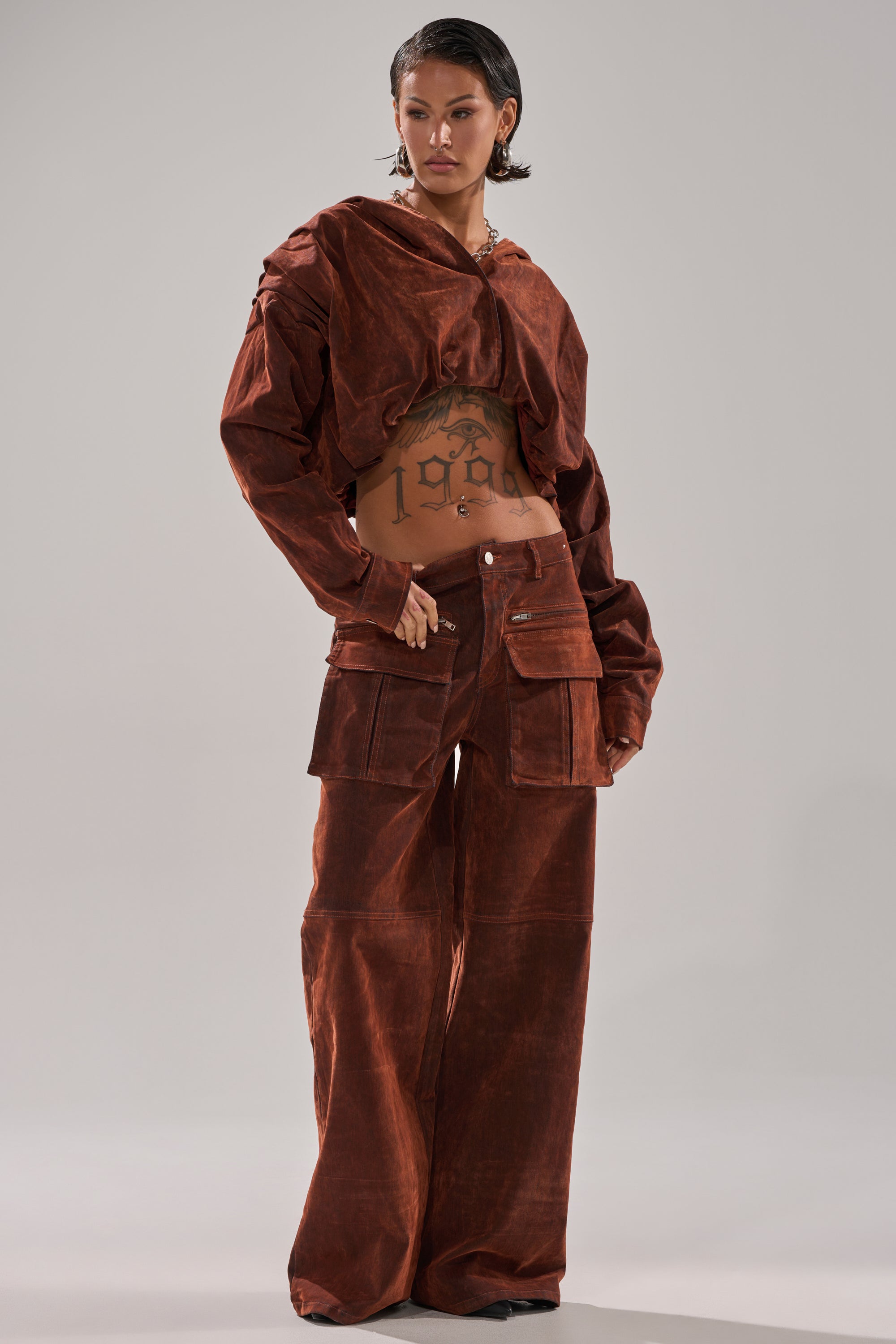 A person with short hair poses in the THINK ABOUT YOU ALL THE TIME VELVET DENIM CARGO TROUSER, rust-colored and wide-leg, paired with a matching oversized jacket, revealing a "1994" tattoo on their torso against a light gray backdrop.