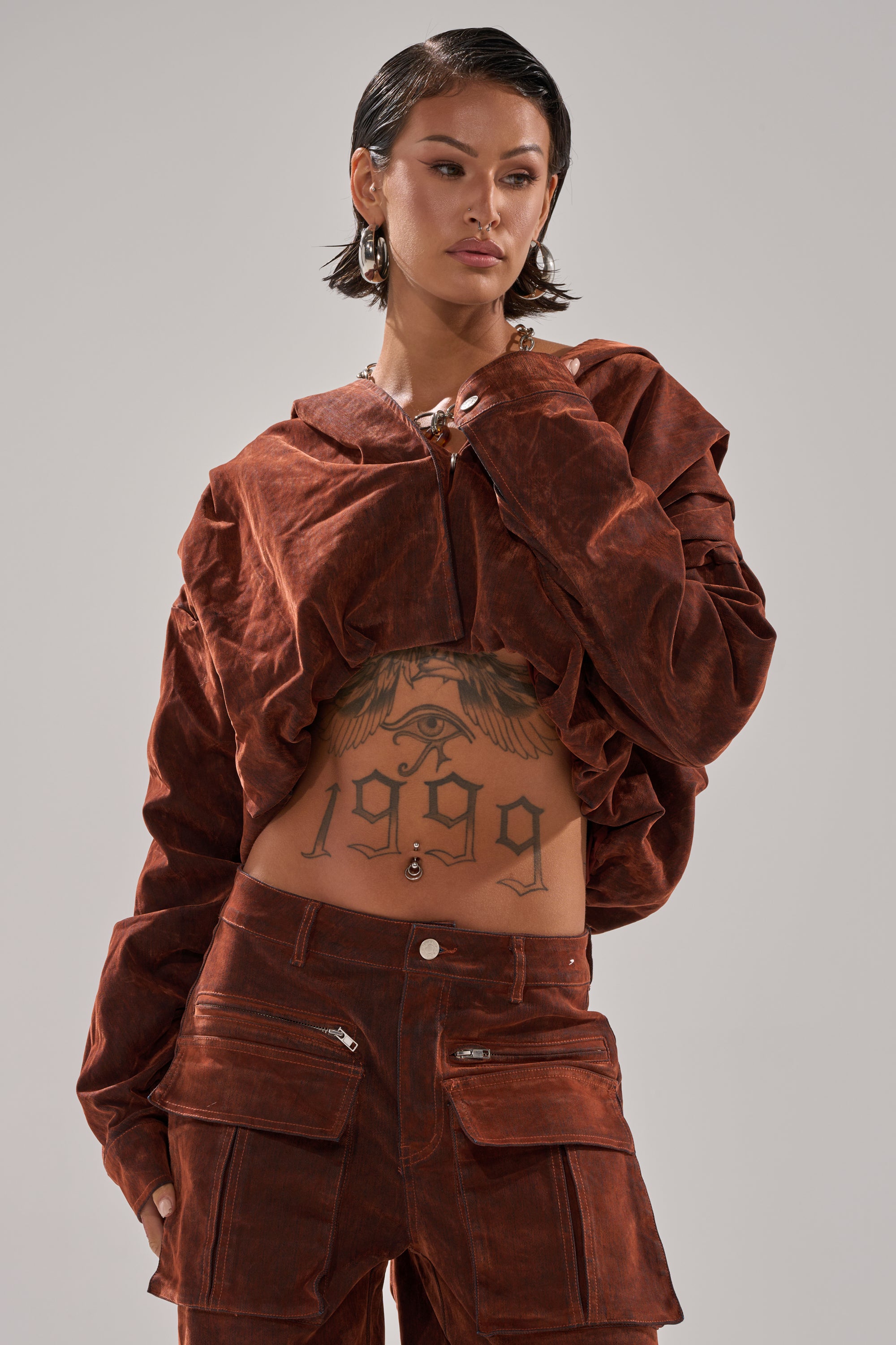 A person with short dark hair wears the THINK ABOUT YOU ALL THE TIME VELVET DENIM LONG SLEEVE TOP and matching pants, revealing a large “1959” eagle tattoo on their stomach, plus earrings and a navel piercing.