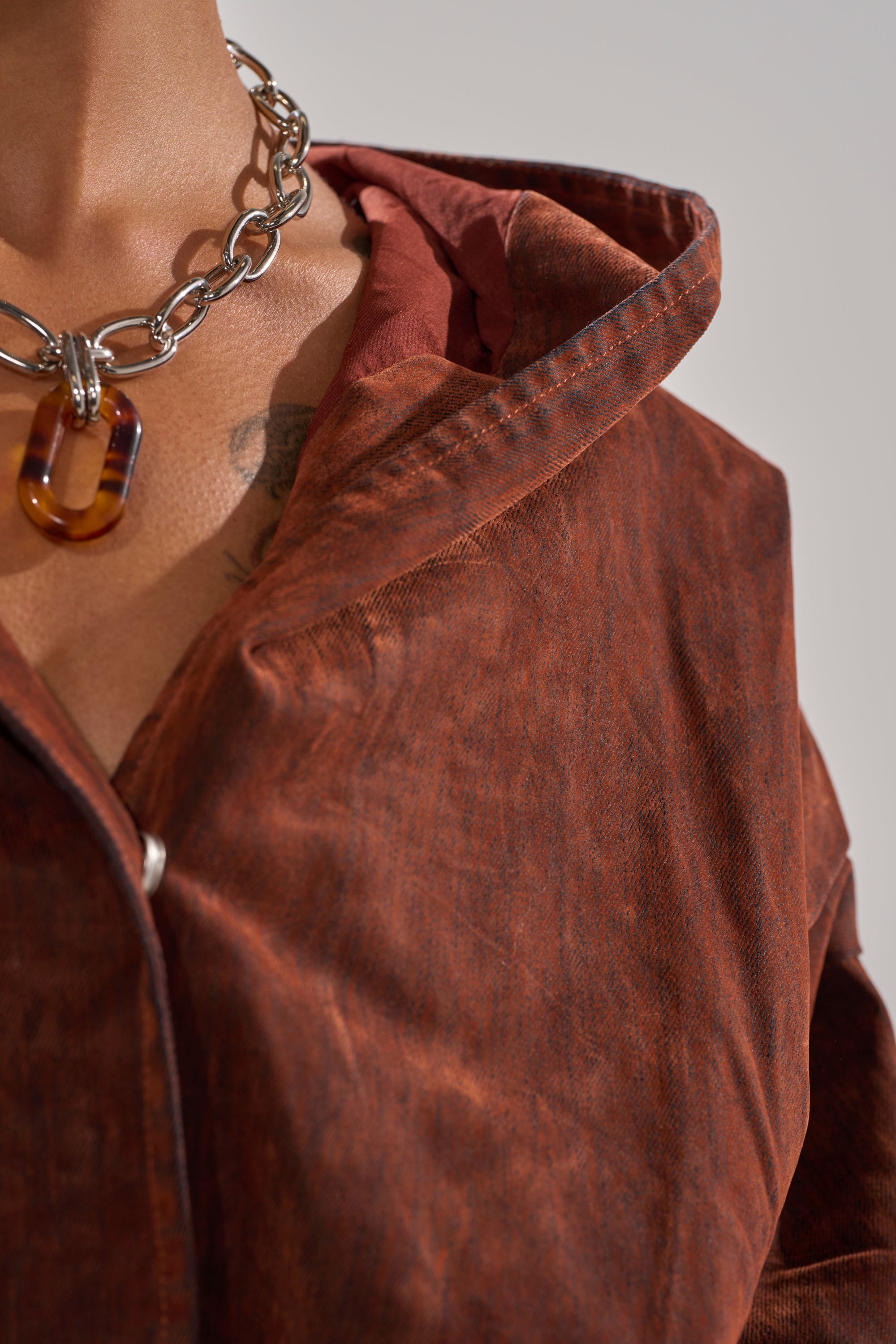 Close-up of a person wearing the THINK ABOUT YOU ALL THE TIME VELVET DENIM LONG SLEEVE TOP in burnt orange over a red shirt, accessorized with a chunky silver chain necklace and brown translucent pendant. A chest tattoo is partially visible.