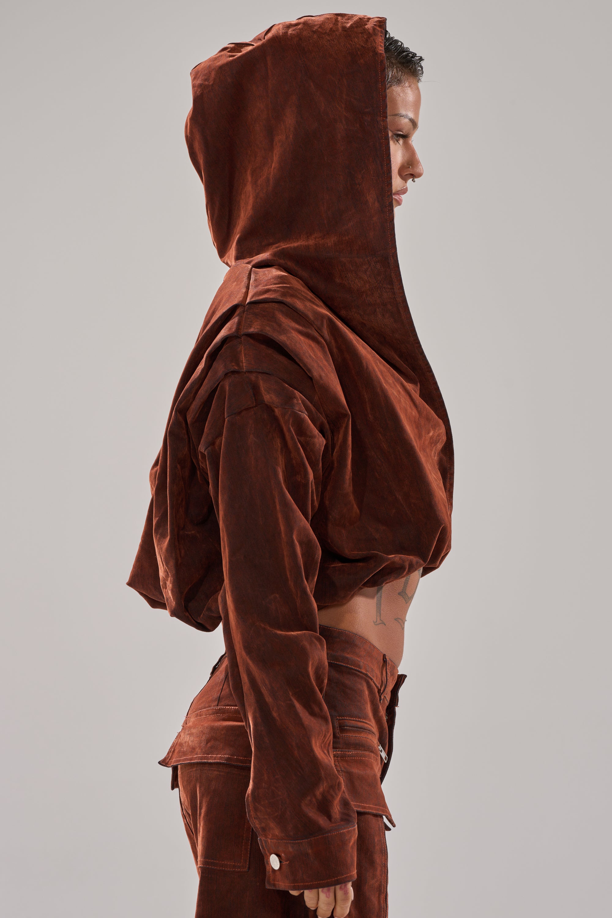 A person in profile wears the THINK ABOUT YOU ALL THE TIME VELVET DENIM LONG SLEEVE TOP in burnt orange with sculptural folds and a suede-like texture, paired with matching pants. The background is plain light gray.