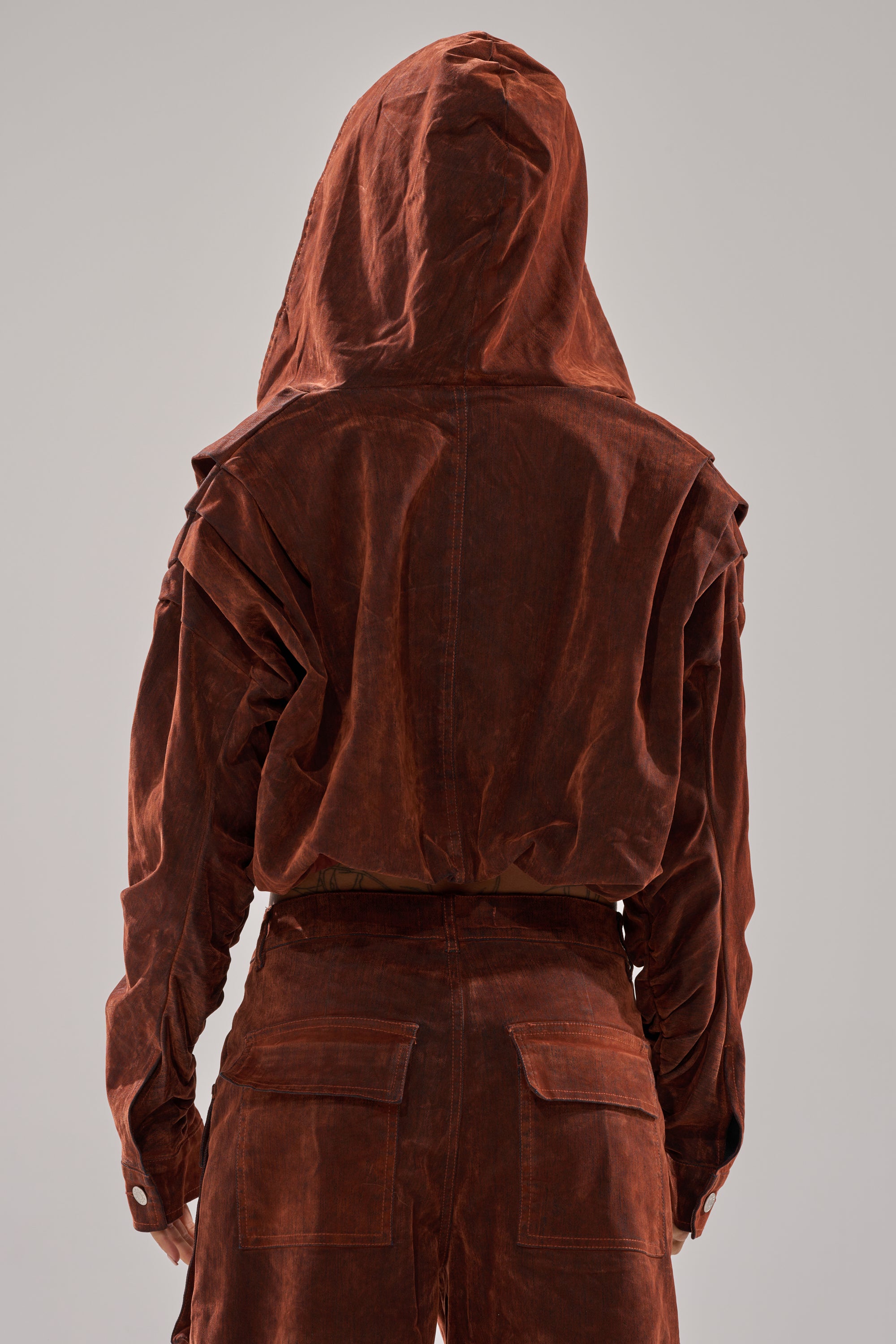 A person is seen from the back wearing the THINK ABOUT YOU ALL THE TIME VELVET DENIM LONG SLEEVE TOP in burnt orange with loose-fitting pants, its slightly wrinkled texture standing out against a plain gray background.