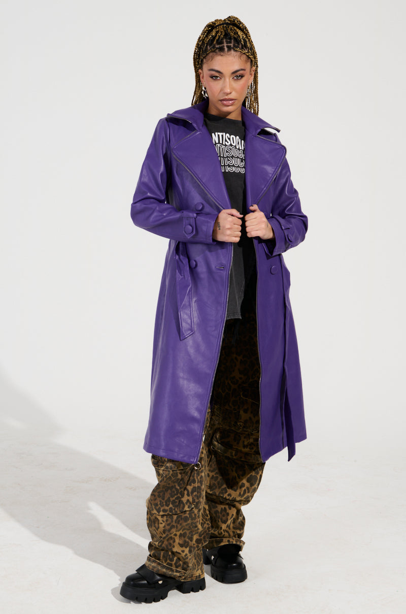 A person confidently stands in a Sando Zipper Trim Trench in purple faux leather over a black graphic T-shirt and leopard-print pants. They have braids and are looking directly at the camera against a plain background.