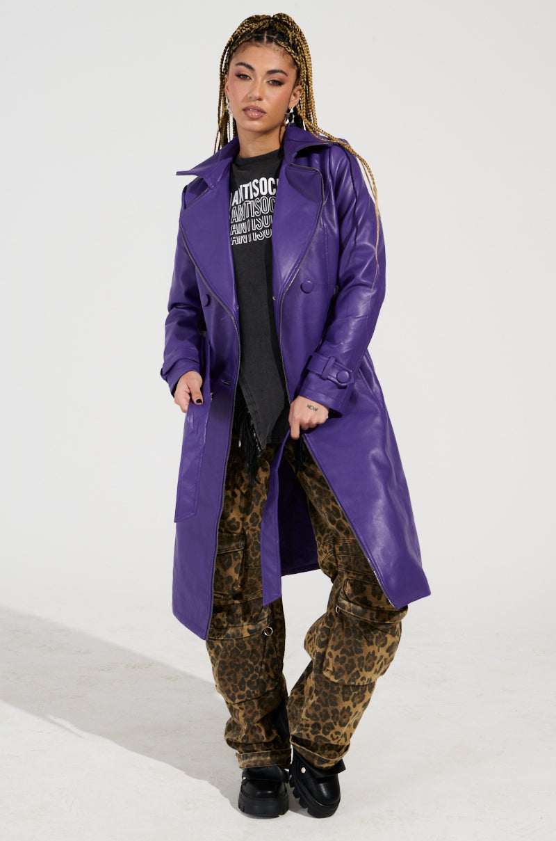 A person stands confidently in the SANDO ZIPPER TRIM TRENCH IN PURPLE, styled over a dark graphic t-shirt and leopard print pants. They have braided hair and are looking at the camera against a plain white background.