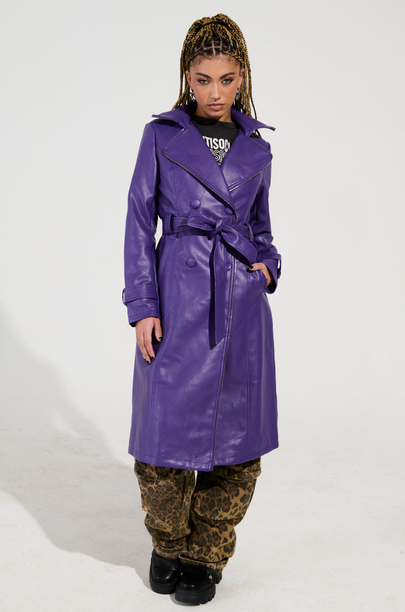 Dressed confidently in the SANDO ZIPPER TRIM TRENCH IN PURPLE with zipper detailing, a person stands on a plain white background. They pair the trench with a black graphic tee and leopard print pants, their expression neutral and hands resting at their sides.