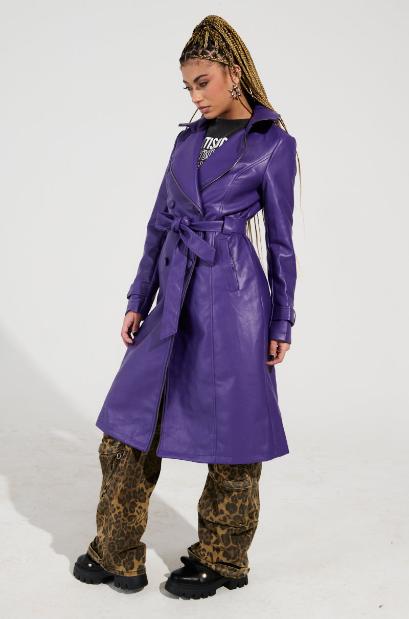 A person with braided hair wears the SANDO ZIPPER TRIM TRENCH IN PURPLE over a black shirt and leopard print pants. They stand against a plain background, looking down to their side with hands by their waist.