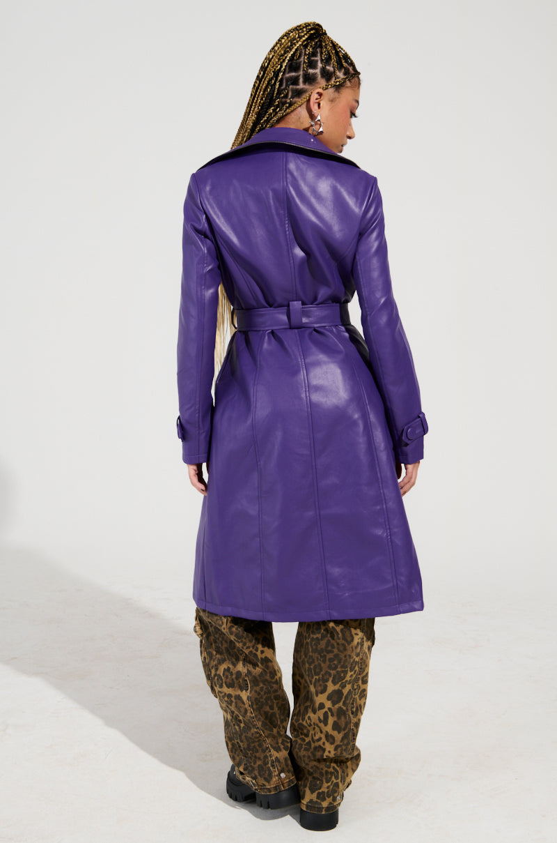 A person with long braided hair is wearing the SANDO ZIPPER TRIM TRENCH IN PURPLE, featuring a waist-cinching belt tied at the back. They complement the look with leopard print pants and black shoes, standing on a plain background.
