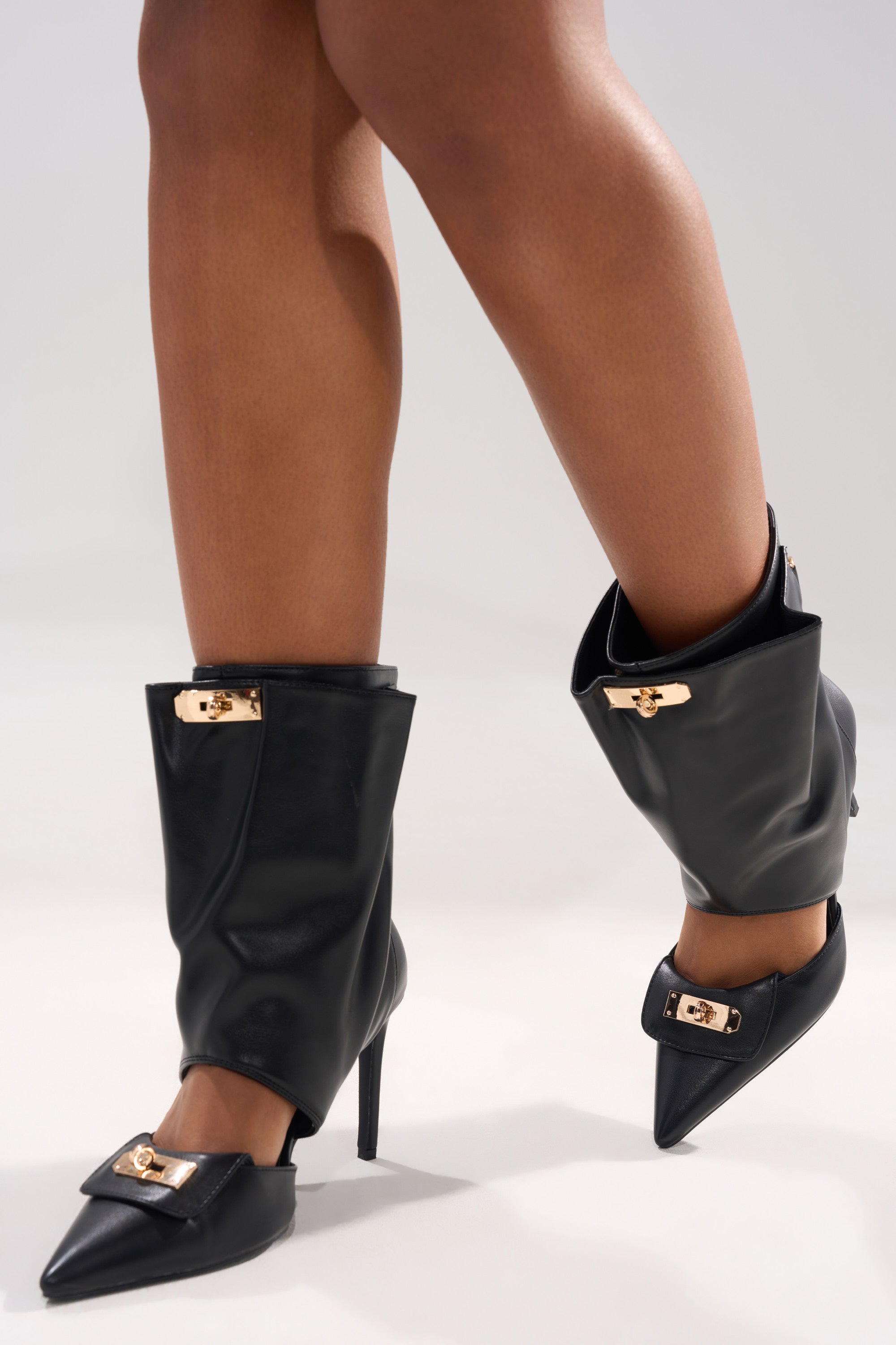A close-up of a person wearing the AZALEA WANG SARVESH BLACK CUTOUT STILETTO BOOTIE, featuring black faux leather, gold buckle accents, pointed toes, and a unique folded-over ankle design. The background is plain and light-colored.