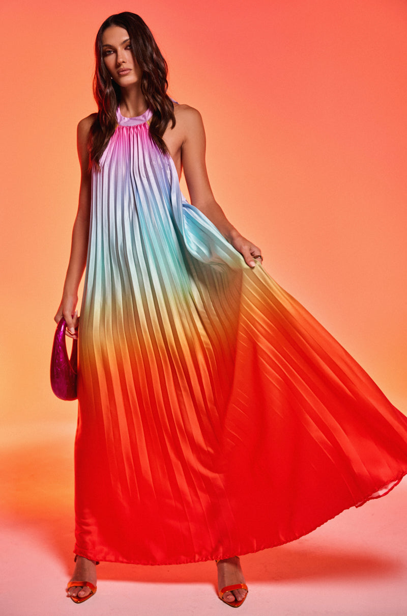 Against an orange backdrop, a woman wears the SUNSET PLEATED OMBRE MAXI DRESS with a halter neckline and blue-to-red rainbow gradient. She displays the pleated satin fabric and holds a small pink purse.