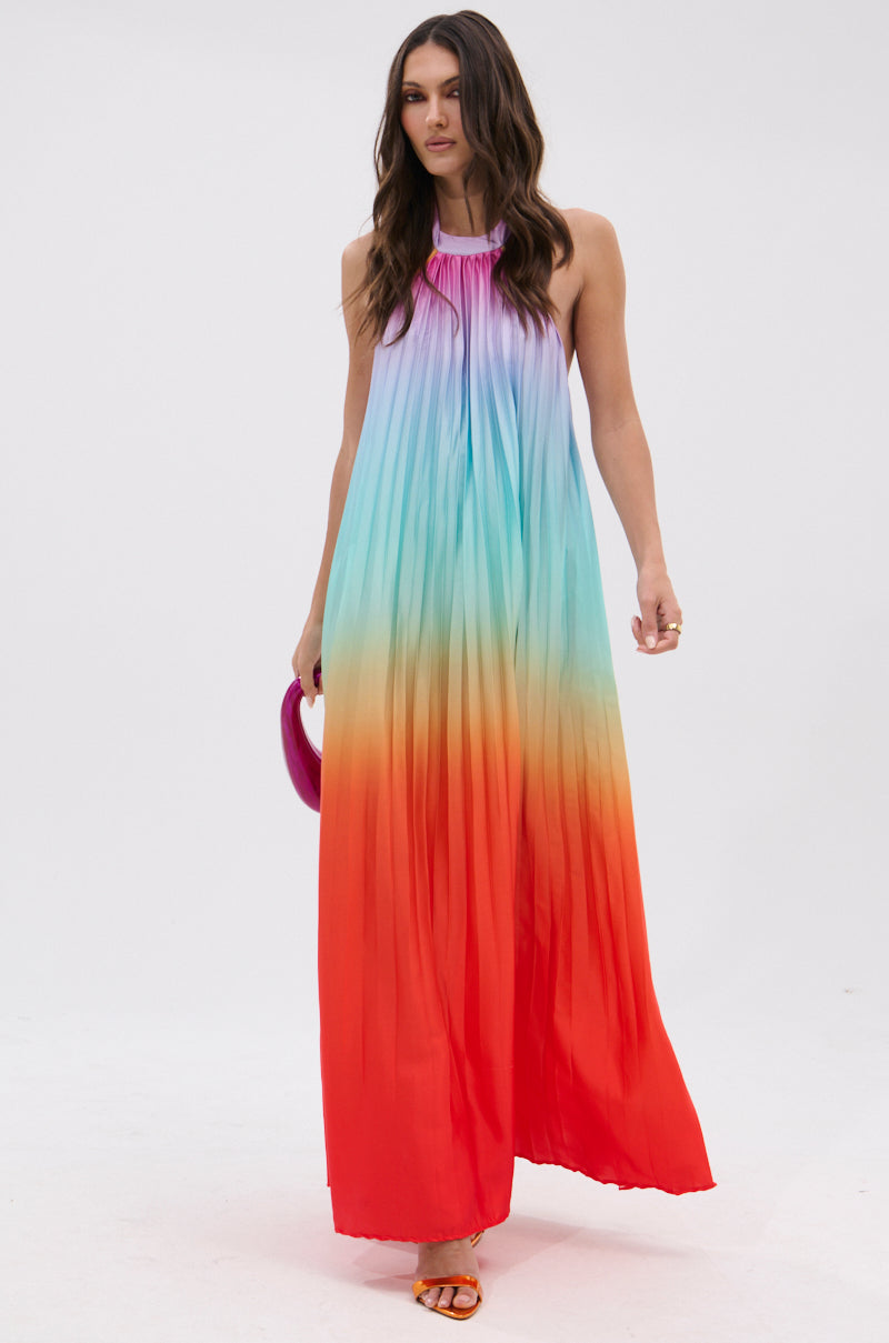 A woman wears the SUNSET PLEATED OMBRE MAXI DRESS, a sleeveless, floor-length gown with a rainbow gradient. She accessorizes with a small purple bag and orange sandals, standing against a plain white background.