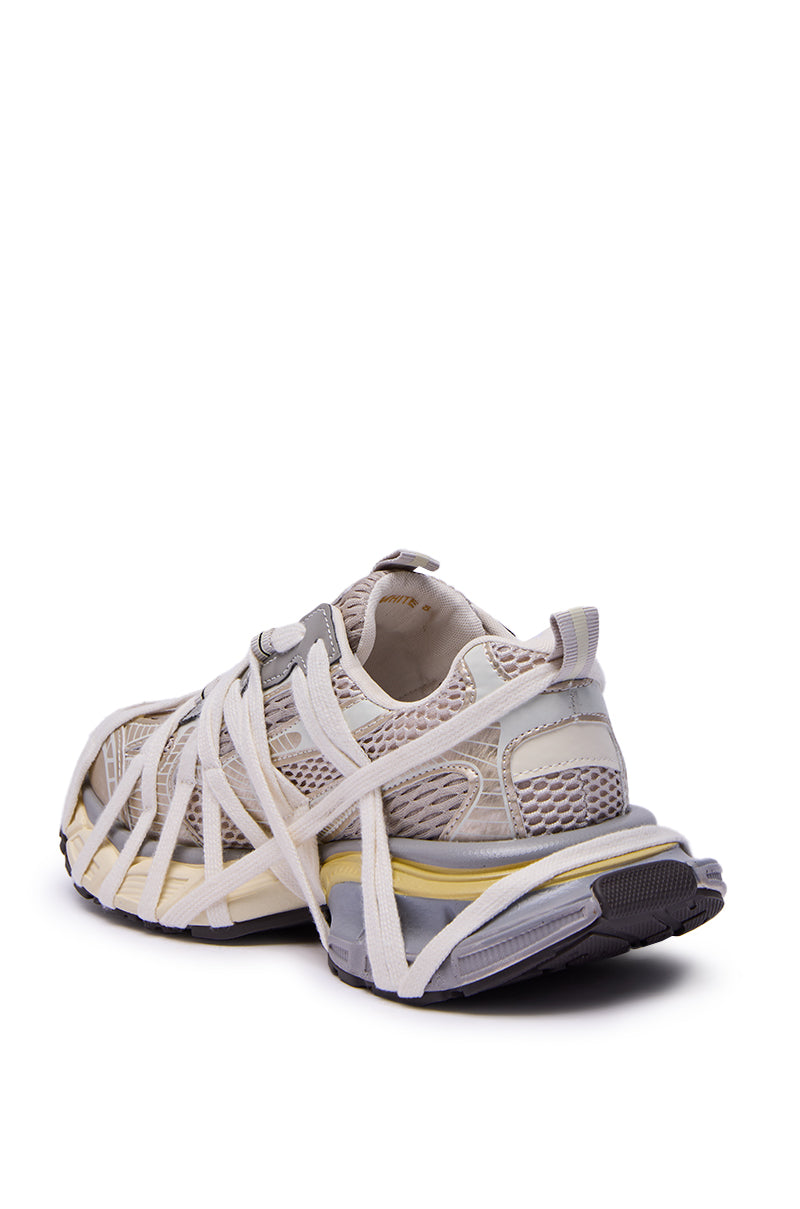 The AZALEA WANG SELIE WHITE FLAT SNEAKER features beige and gray mesh panels, white crisscross elastic straps, and a chunky sole with yellow accents. Shown at an angle from the back and side.