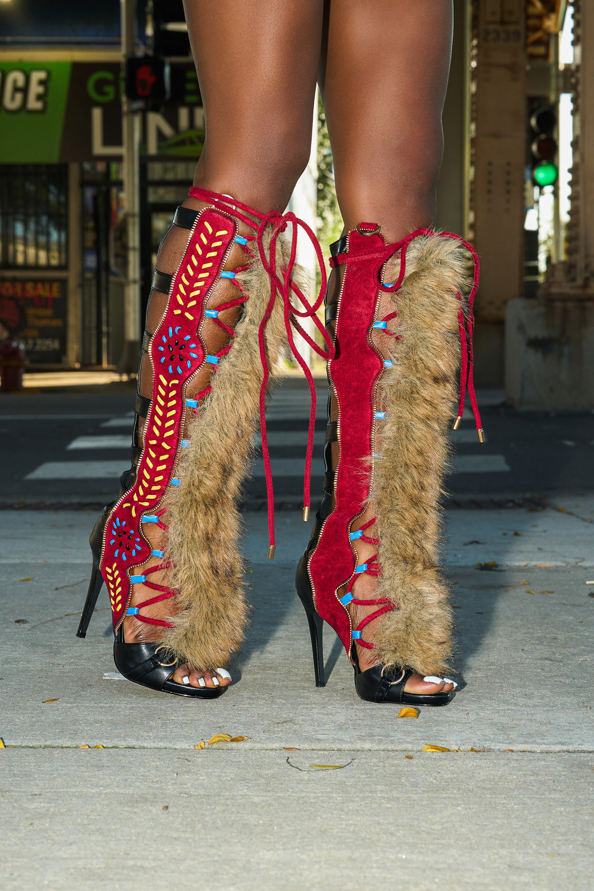 A person stands on a city sidewalk in AZALEA WANG SHAINE BLACK MULTI GLADIATOR STITCH AND FUR SANDALS, featuring high heels, red suede with faux fur trim, colorful beadwork, lace-up details, and open toes. Urban street and storefronts form the background.