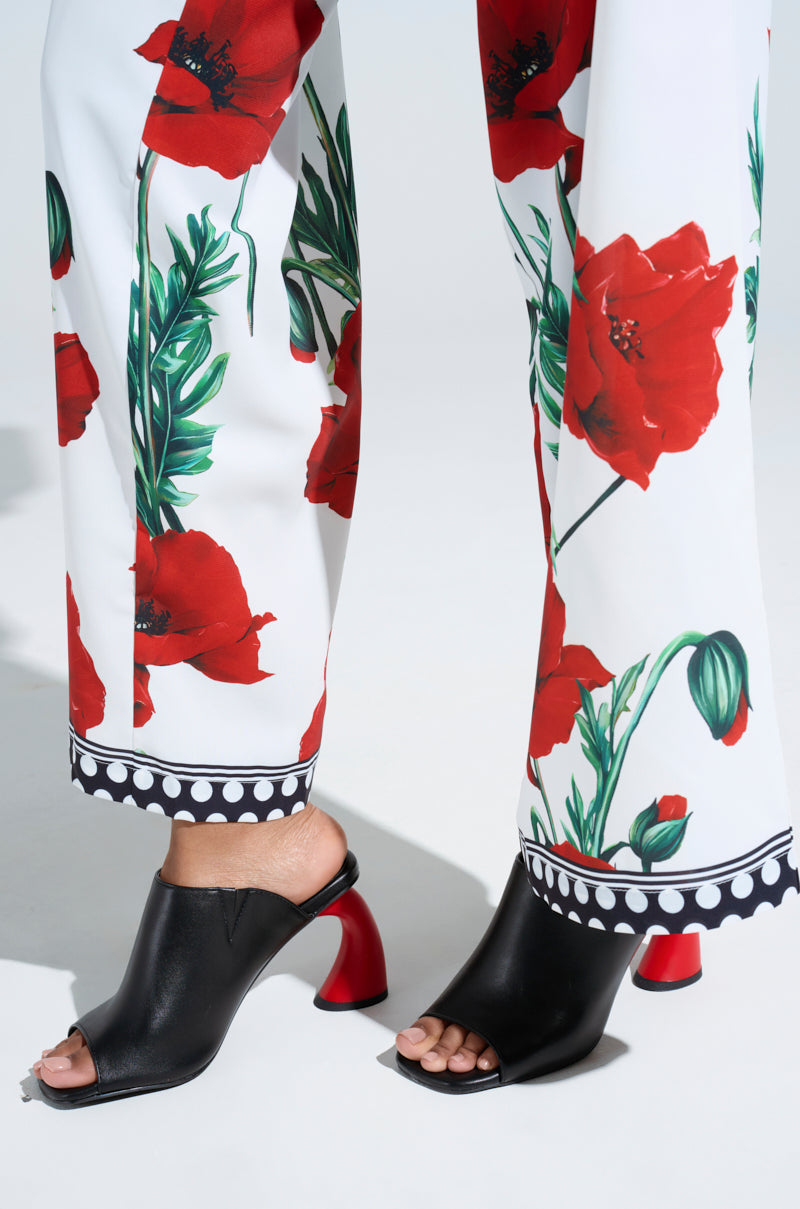 The close-up features a femme fatale look: white pants with a vibrant red poppy print and dotted border, paired with the AZALEA WANG SIDLE black slip-on mule sandals accented by bold, red curved heels for an effortlessly chic ensemble.