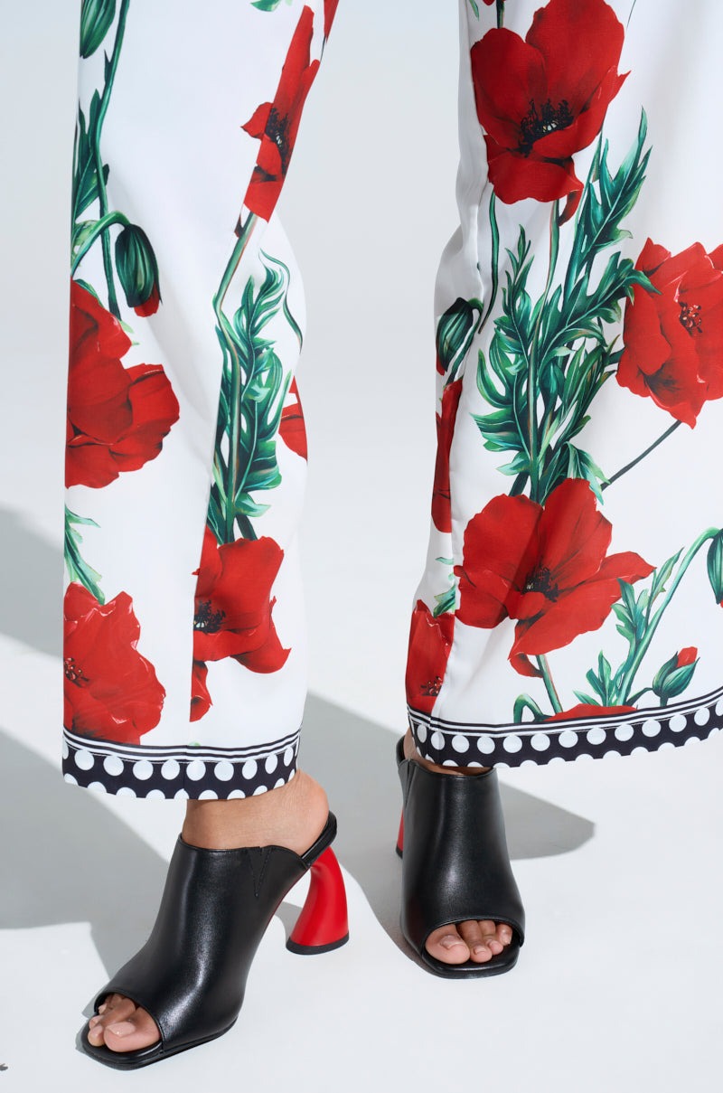 Embodying a femme fatale vibe, the person wears white pants with red poppies and chic AZALEA WANG SIDLE BLACK SLIP ON MULES. These sandals feature an open toe and striking red chunky heels set against a neutral, light-colored background.