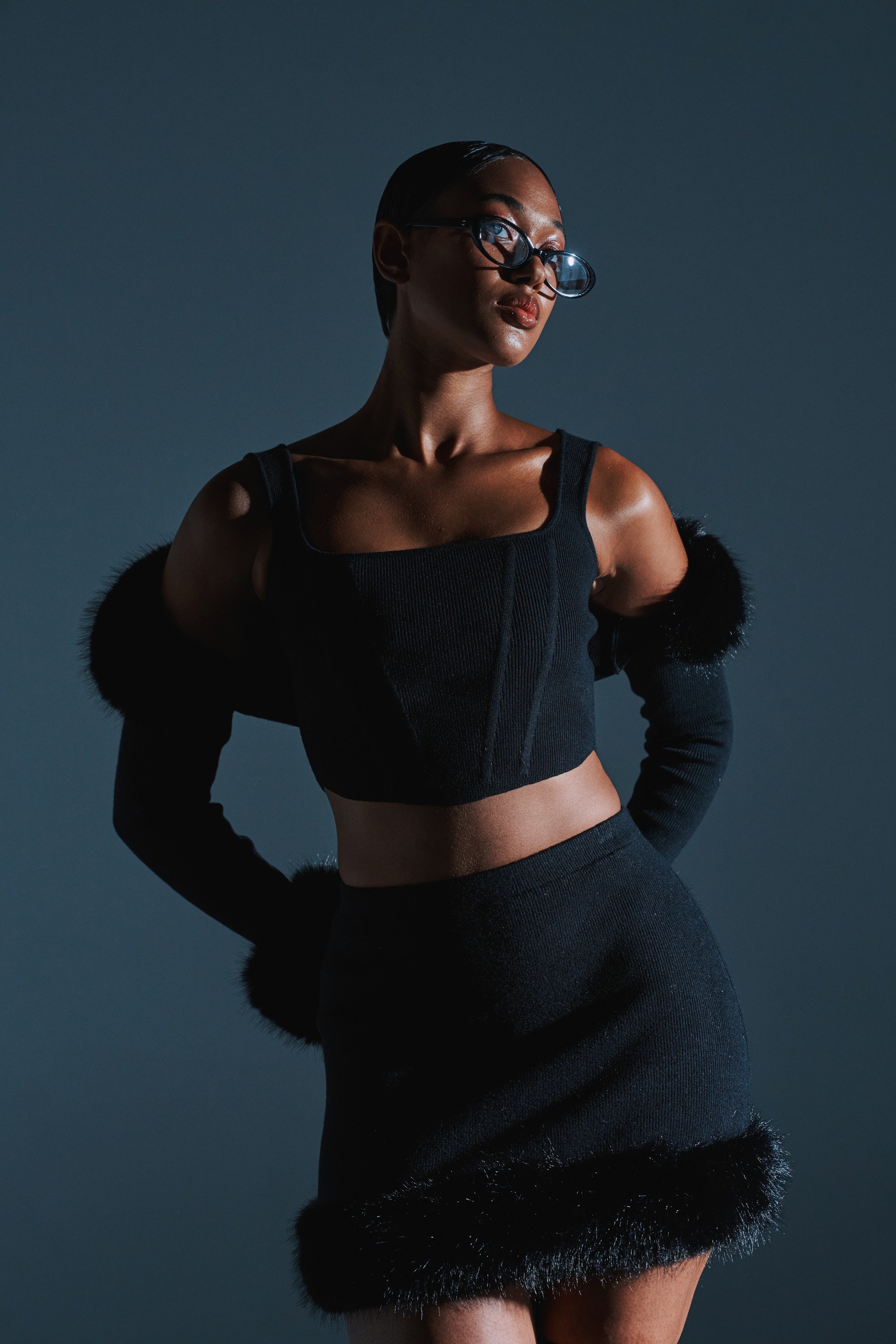 A woman poses confidently against a dark background, wearing the BIG MOOD FITTED CORSET IN BLACK styled with a matching skirt, long black gloves, faux fur trim, and fashionable glasses.