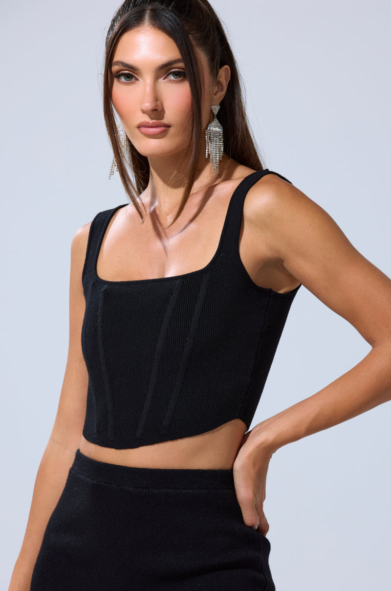 A woman with long brown hair and large earrings poses in the BIG MOOD FITTED CORSET IN BLACK, a sleeveless, square-neck ribbed knit crop top, paired with matching high-waisted pants against a plain light background.