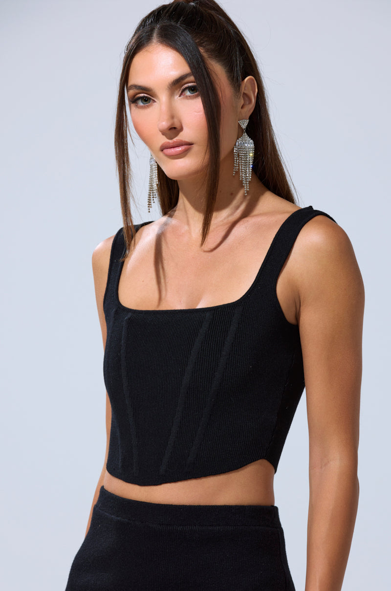 A woman with long brown hair confidently wears the BIG MOOD FITTED CORSET IN BLACK and statement silver earrings, posing against a plain light gray background.