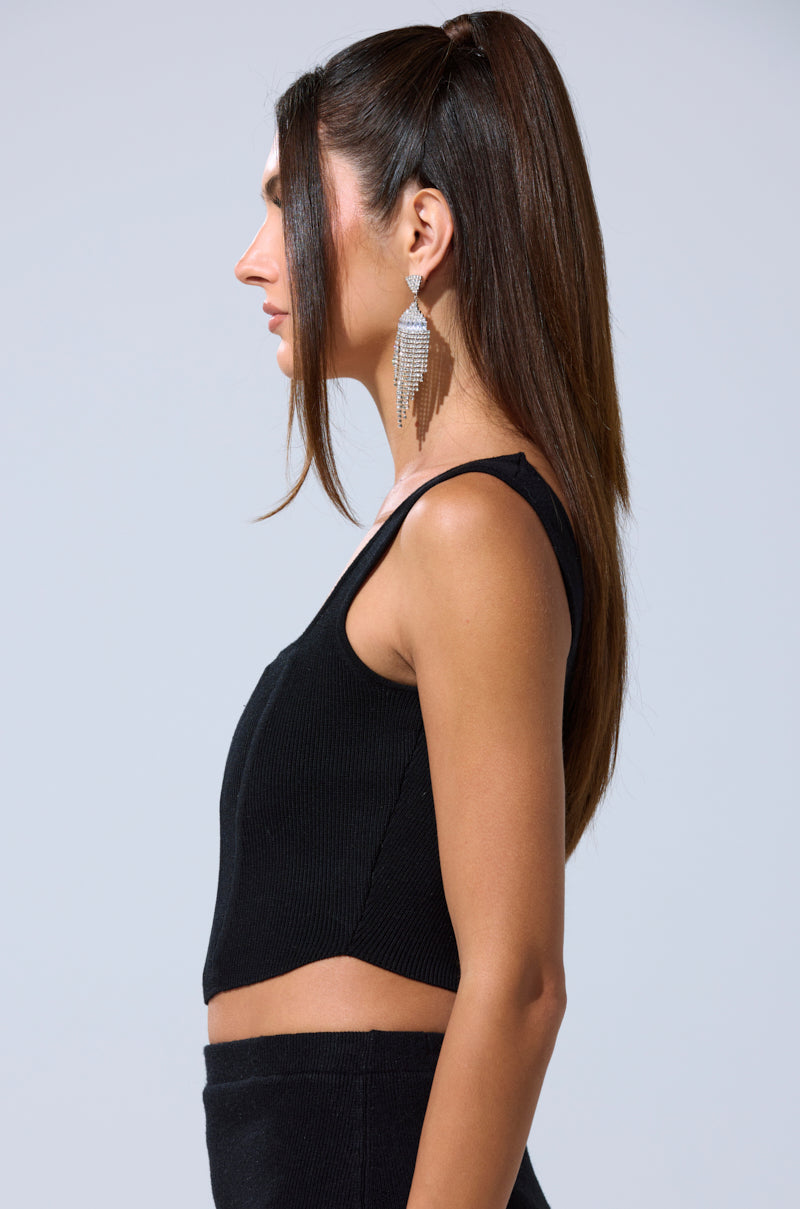 A woman with long straight brown hair in a high ponytail, wearing the BIG MOOD FITTED CORSET IN BLACK and large dangling earrings, stands in profile against a plain light gray background.