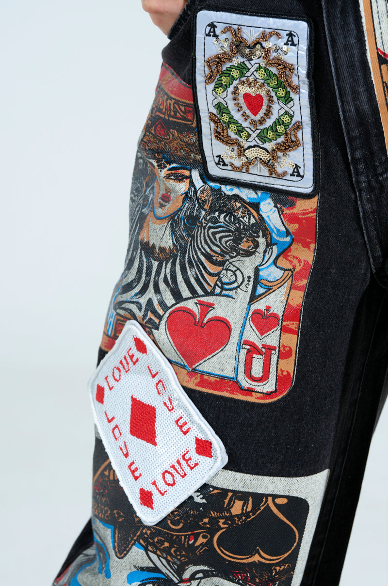 Close-up of black embellished Queen of Hearts Printed and Embellished Denim Pants with vibrant playing card graphics, including aces and hearts, plus a "LOVE" patch with a red diamond.