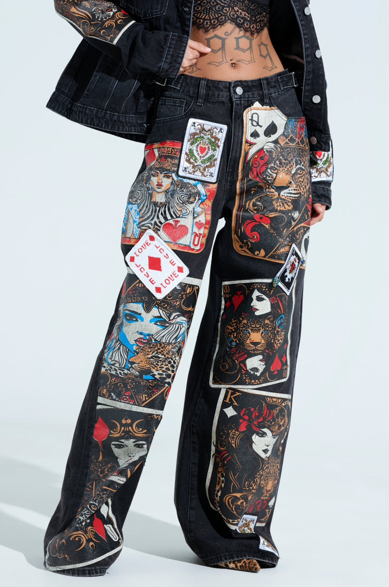 A person in a black denim outfit stands out with Queen Of Hearts Printed and Embellished Denim Pants, featuring playing card graphics, artistic faces, vibrant motifs, and a top adorned with patterns of love symbols, a crown, and intricate details.