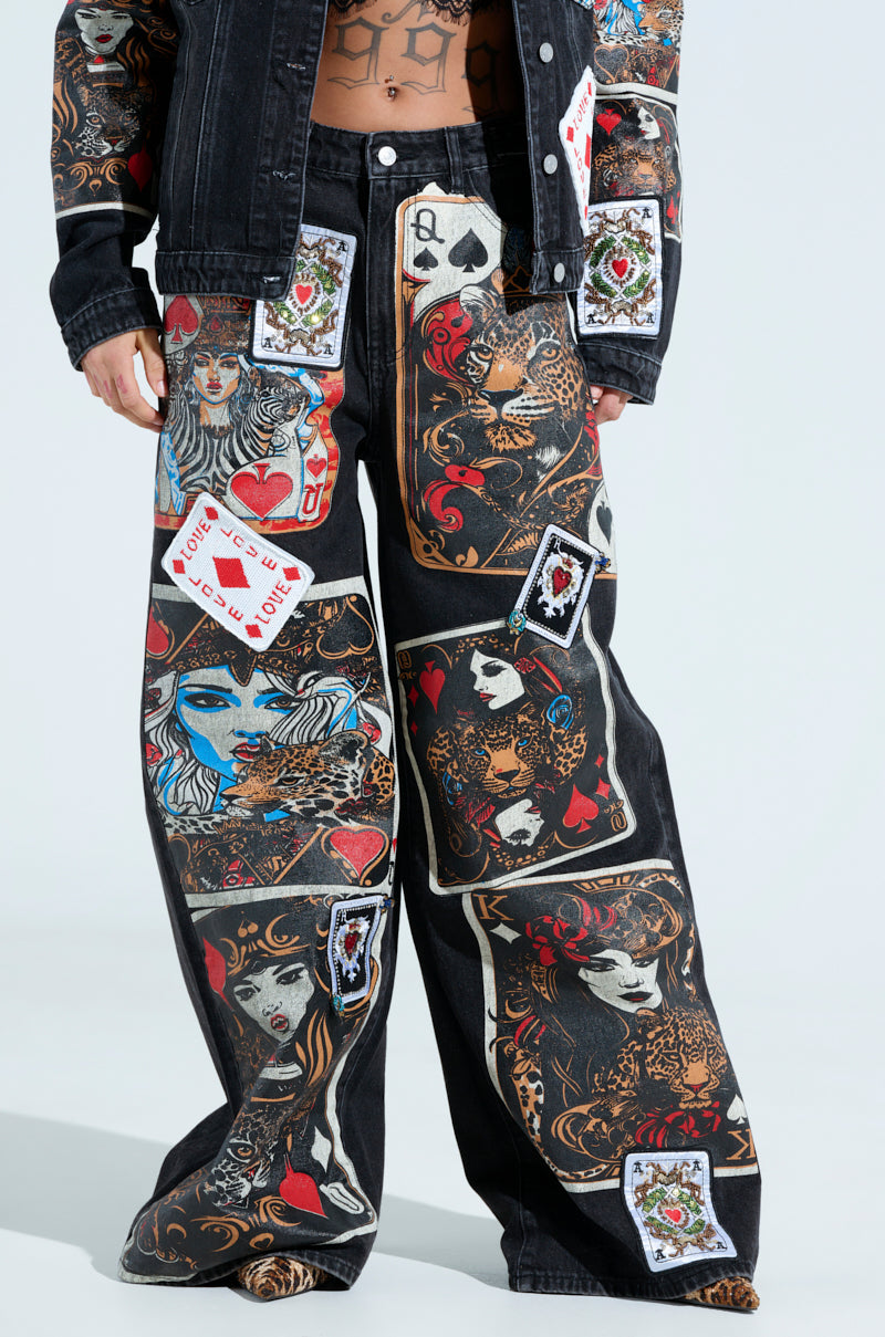 Wearing a black denim jacket and wide-leg jeans, someone sports Queen Of Hearts Printed and Embellished Denim Pants, showcasing vibrant playing card graphics—kings, queens, hearts—in red and blue. The outfit also features intricate leopard and floral designs for an artistic flair.