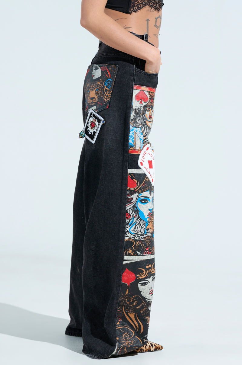 A person models the QUEEN OF HEARTS PRINTED AND EMBELLISHED DENIM PANTS adorned with vivid playing card graphics, featuring faces and symbols. The denim is enhanced by colorful patches and paired with a cropped black lace top.