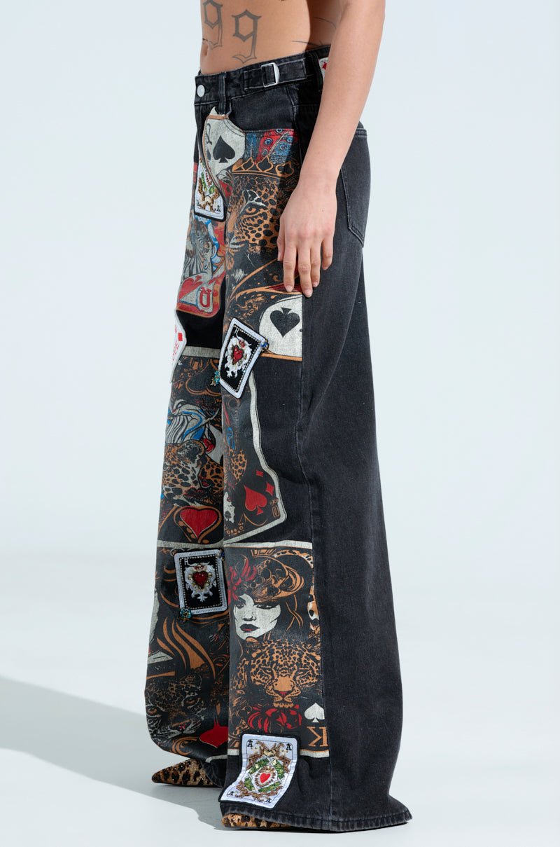 A person wearing QUEEN OF HEARTS PRINTED AND EMBELLISHED DENIM PANTS featuring colorful patchwork with card graphics, including the queen, and abstract art. These relaxed-fit, high-waist jeans stand out against a plain light background.