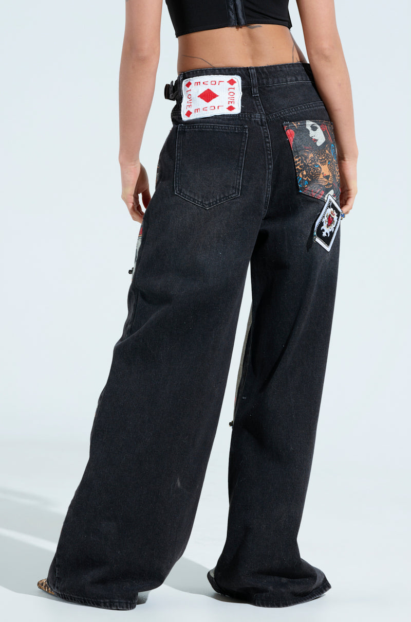 Rear view of a person in QUEEN OF HEARTS PRINTED AND EMBELLISHED DENIM PANTS, featuring wide-legged black jeans with playing card graphics and patches. Paired with a black cropped top against a plain light background.