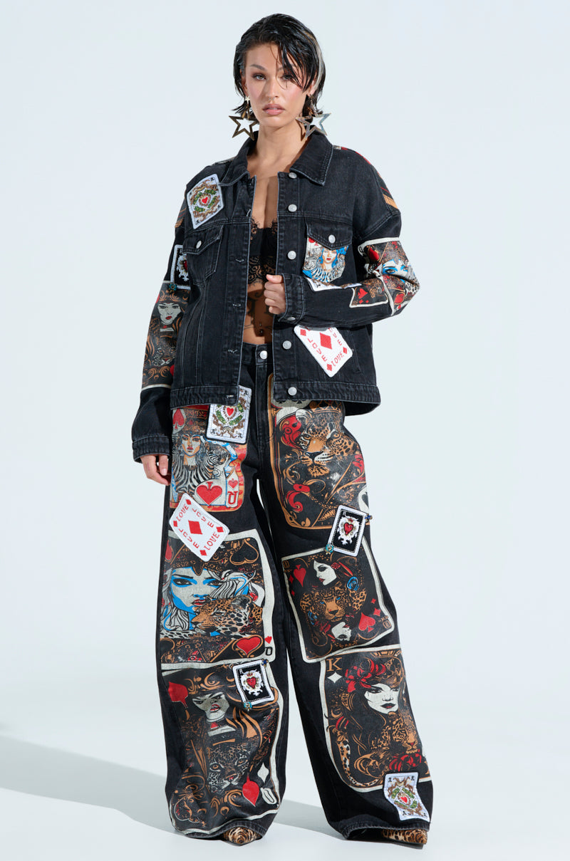 Person posing in a black denim jacket and QUEEN OF HEARTS PRINTED AND EMBELLISHED DENIM PANTS, adorned with vibrant card-themed patches, graphics, and motifs like faces, hearts, and spades on a light gray background.