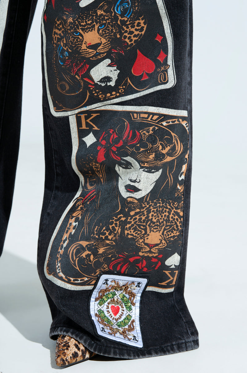 Close-up of QUEEN OF HEARTS PRINTED AND EMBELLISHED DENIM PANTS shows black pants adorned with vivid playing card graphics, featuring a woman with a red rose and a leopard. Additional heart and spade motifs add artistic flair, hinting at Queen Of Hearts themes.