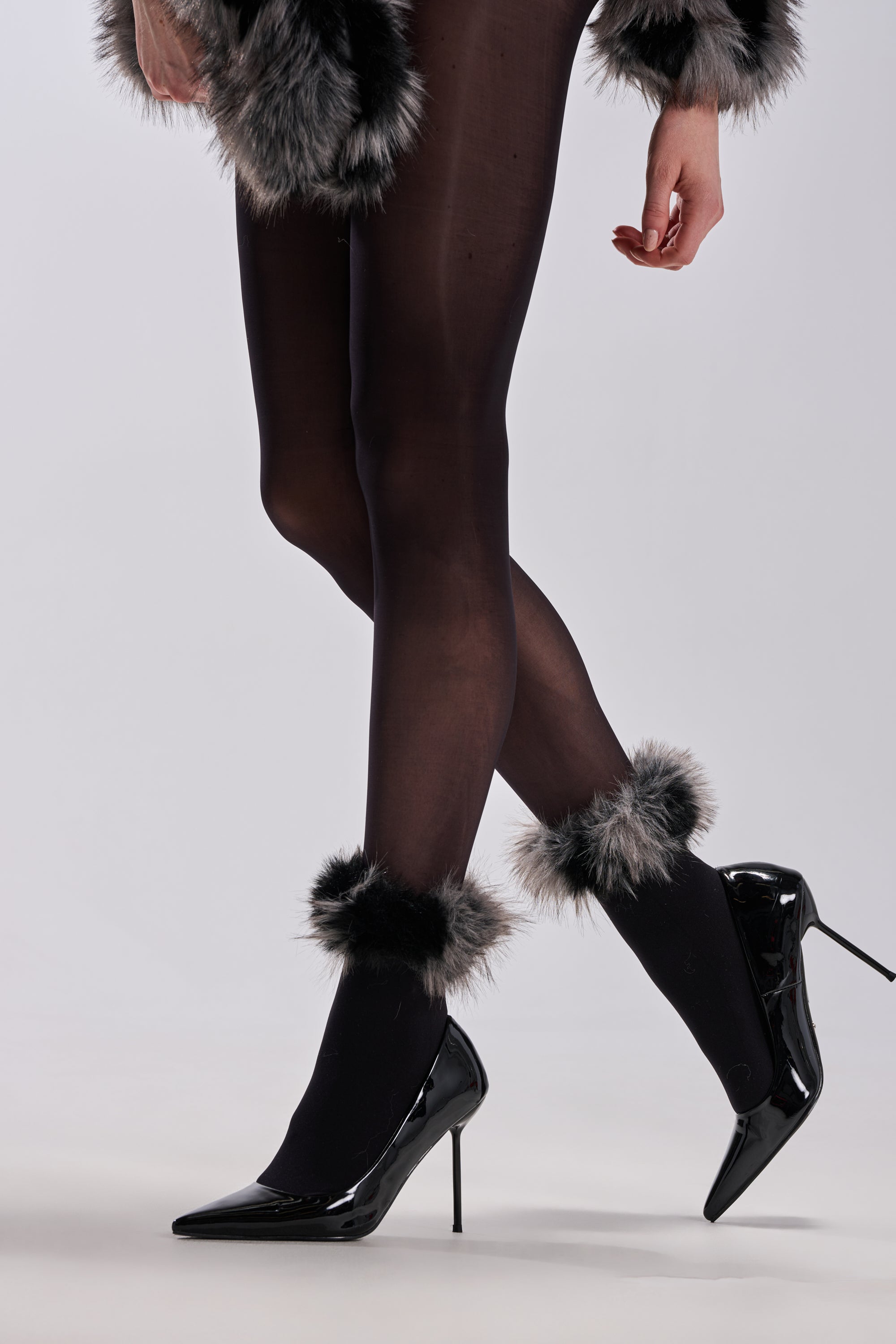 A person stands against a neutral background wearing black sheer tights, black patent high heels, and ARLO FAUX FUR TRIM SOCKS featuring cozy chic gray faux fur on the cuffs and anklets.