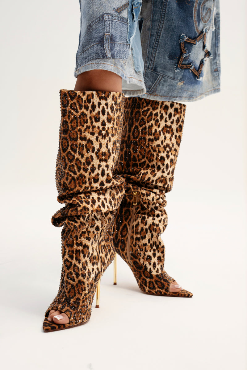 A person flaunts the AZALEA WANG SPHYNX LEOPARD OPEN TOE BOOT, knee-length with high stiletto heels and subtle rhinestone embellishments, paired perfectly with blue distressed jeans rolled up at the bottom.