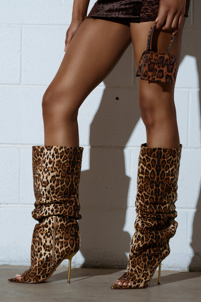 Wearing AZALEA WANG SPHYNX leopard open toe boots, a person stands against a white wall with a textured handbag. Paired with a brown mini skirt, the ensemble creates a stylish and bold fashion statement.