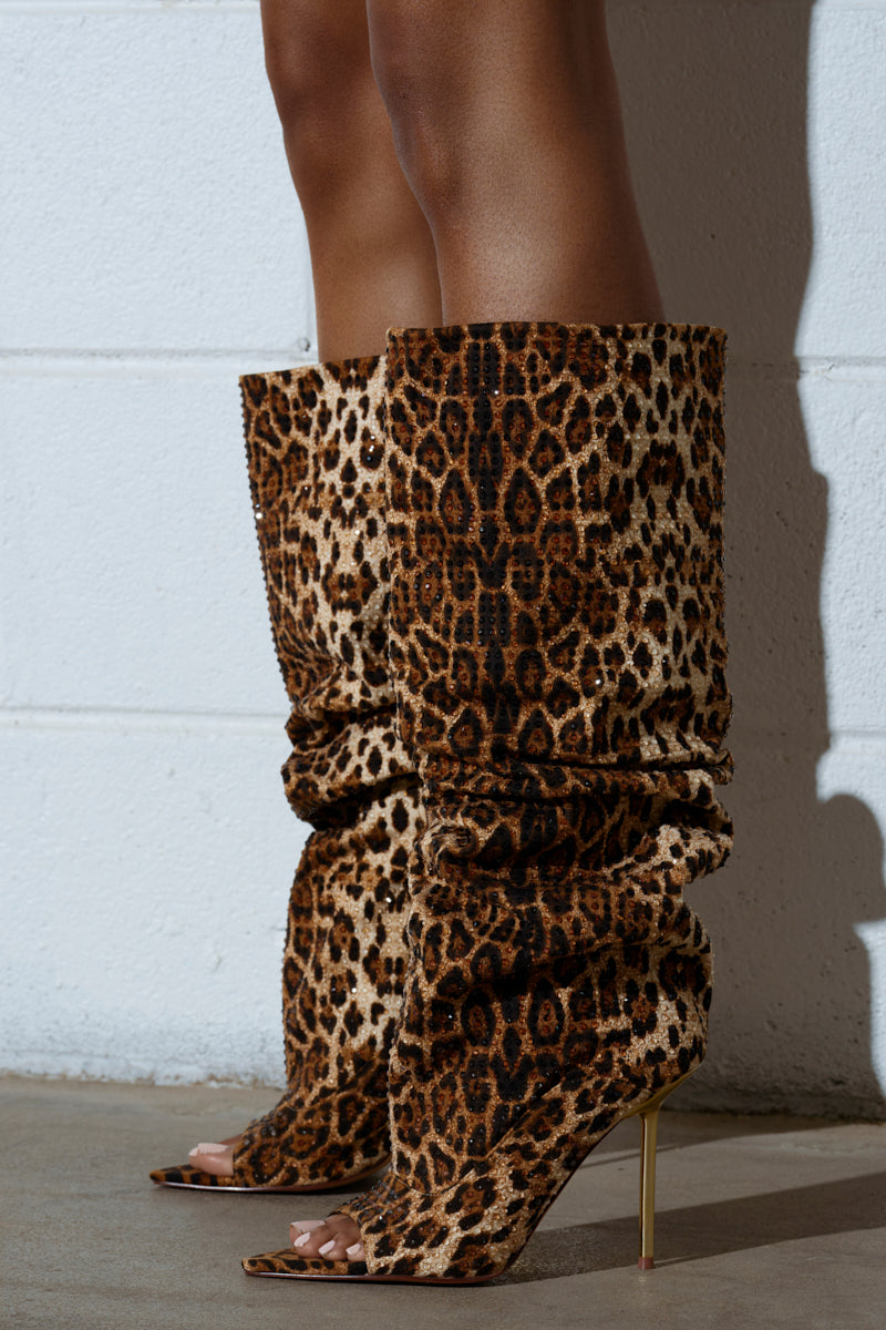 A person confidently stands on a concrete floor against a white brick wall, wearing AZALEA WANG SPHYNX LEOPARD OPEN TOE BOOTS—striking knee-high faux suede stilettos with open toes.