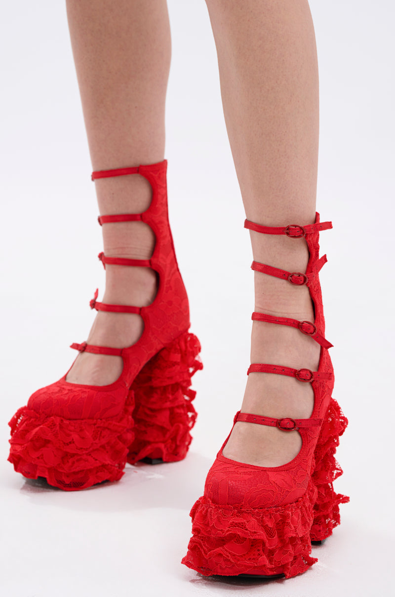 A person wearing the AZALEA WANG SPREENE RED CHUNKY PUMP, featuring bold red faux leather, multiple straps, chunky platform heels, and ruffled details, stands on a plain white background.
