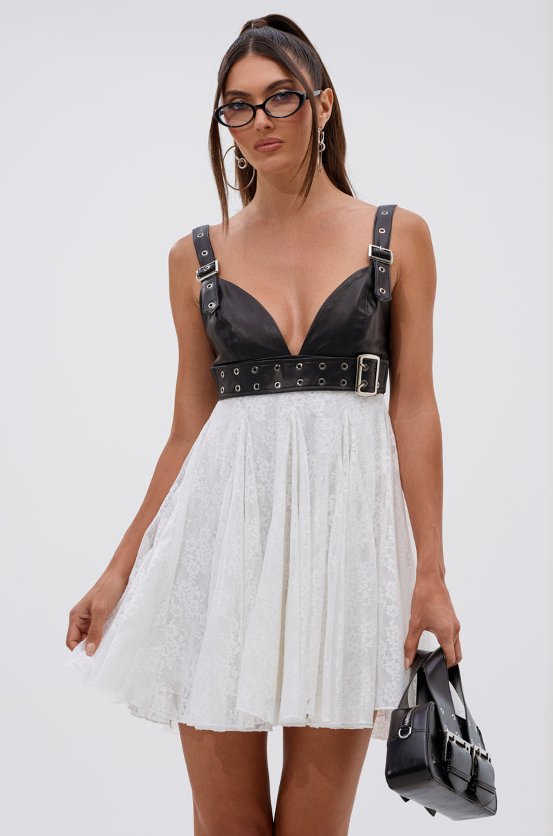 A woman wears the EDGY WITH A BOW ON TOP MINI DRESS, featuring a sleeveless faux leather bodice and white floral lace skirt. She holds a black handbag, accessorized with hoop earrings and cat-eye glasses. The background is plain light gray.