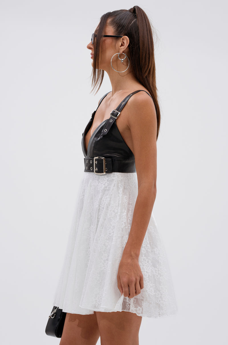A woman with long brown hair in a high ponytail wears sunglasses, hoop earrings, and the EDGY WITH A BOW ON TOP MINI DRESS—featuring a faux leather bodice and white floral lace skirt—while holding a black bag against a plain background.