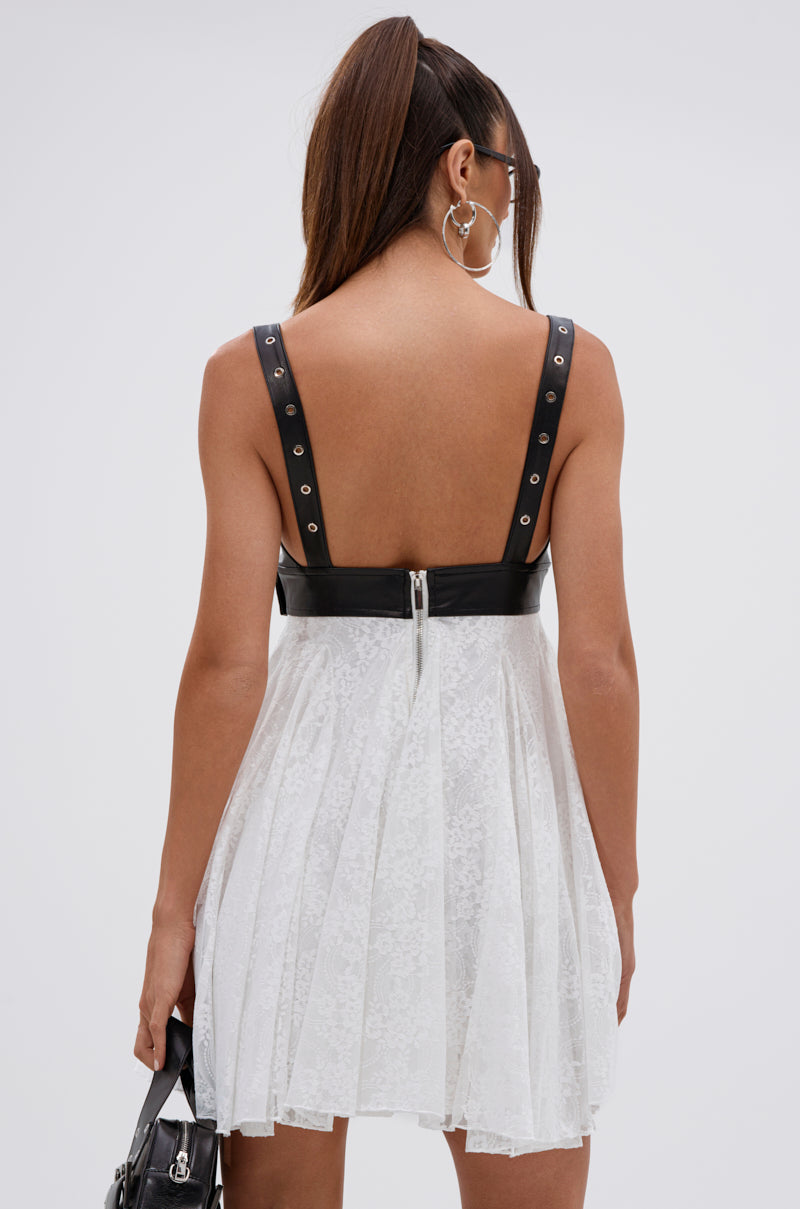 A woman with long brown hair in a high ponytail wears the EDGY WITH A BOW ON TOP MINI DRESS—a white mini dress with a faux leather bodice, silver grommets, floral lace skirt, and back zip—paired with a black handbag.