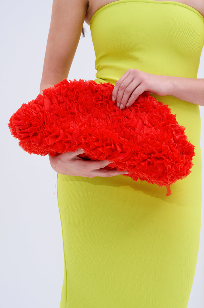 A person in a strapless, bright yellow dress holds the RUFFLED UP CLUTCH—an oversized bag with bold texture—against a plain light background.