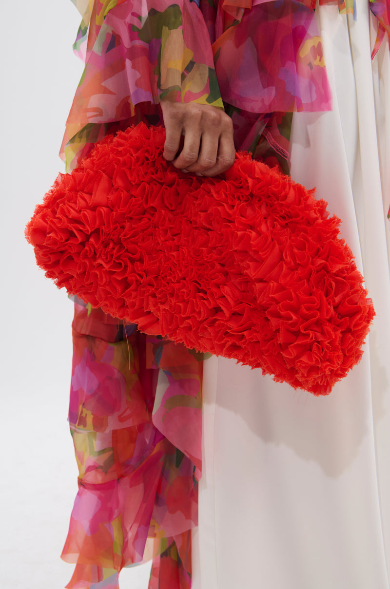 A person in a flowing, colorful floral dress holds the RUFFLED UP CLUTCH—a vibrant red, ruffled statement accessory—while standing against a light background.